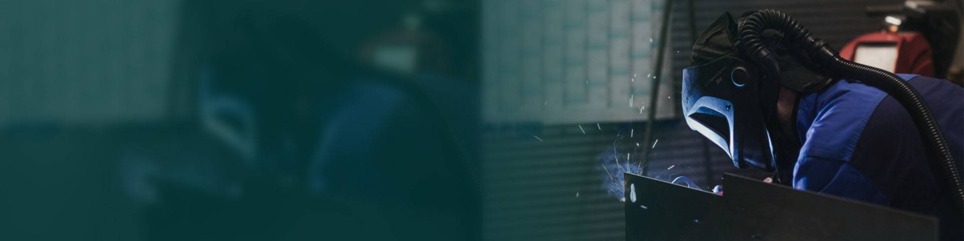 A welder works on a piece of metal in a workshop, wearing a safety helmet and respirator.