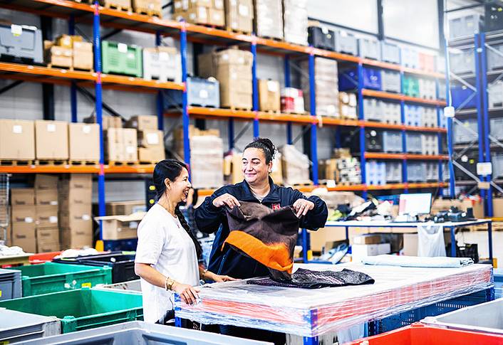 Two women fold clothes in a warehouse full of cardboard boxes.
