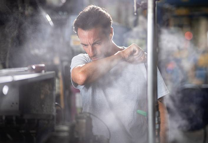 In a smoky industrial environment, a man coughs into his elbow.
