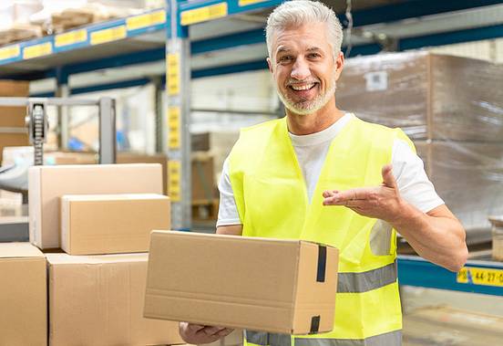 Warehouse worker in high-visibility waistcoat holds a clean parcel in a warehouse and smiles into the camera