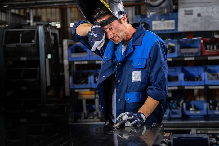 Schweißer steht mit hochgeklapptem Helm in einer Werkstatt und reibt sich das Auge.