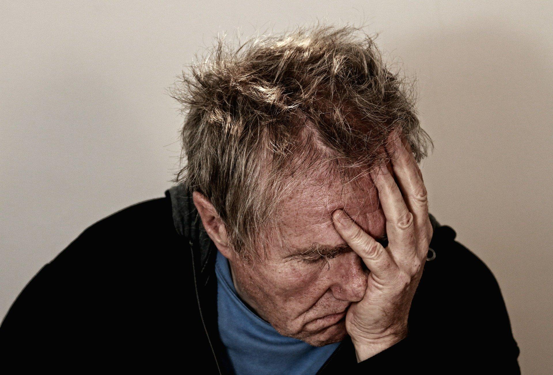 Elderly man with dishevelled hair holds his head in his hand, exhausted and desperate.