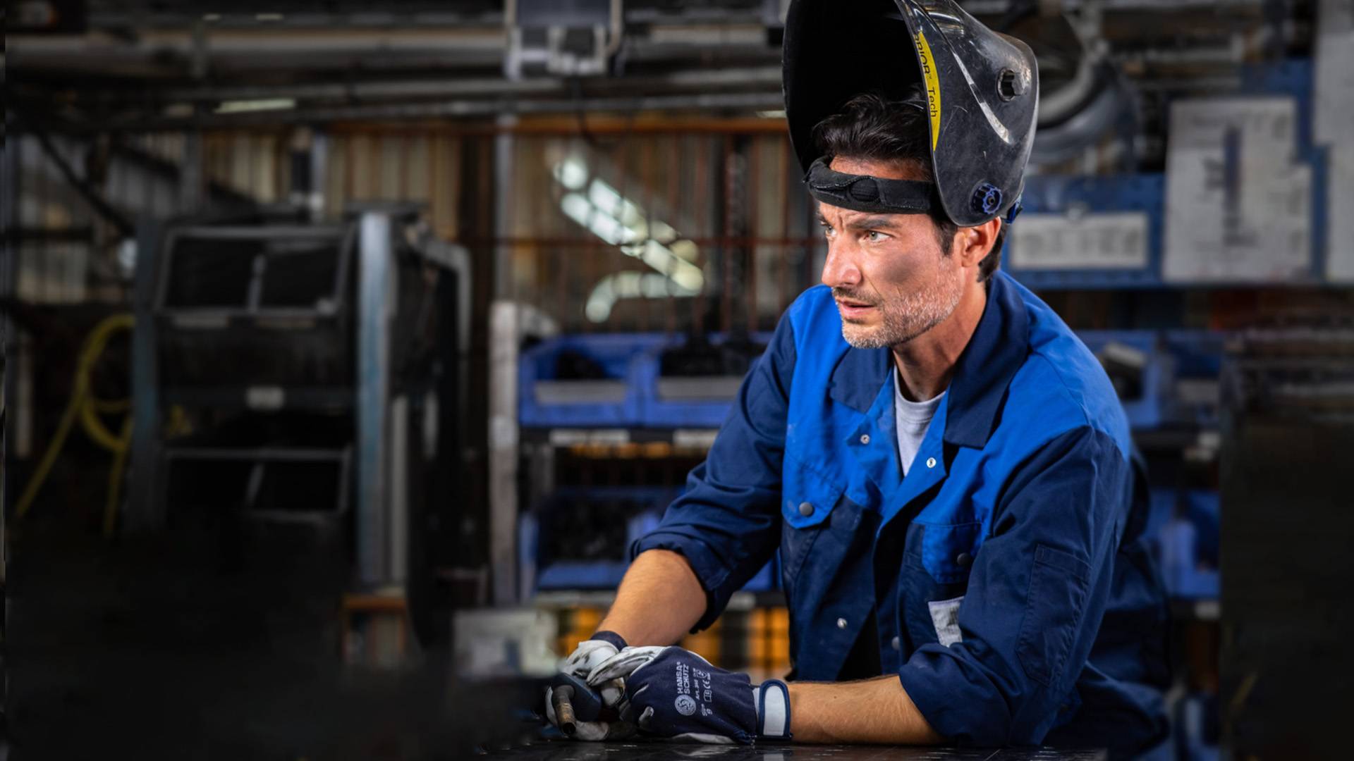 A welder in blue work clothes with his hard hat up is standing in a workshop.