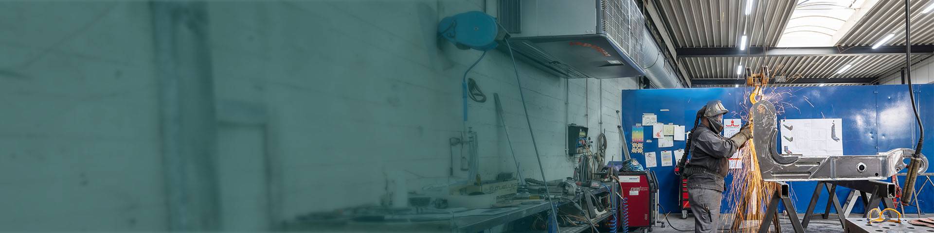 Welder in a metal workshop working on a large component with flying sparks; an industrial air purifier is installed behind him.