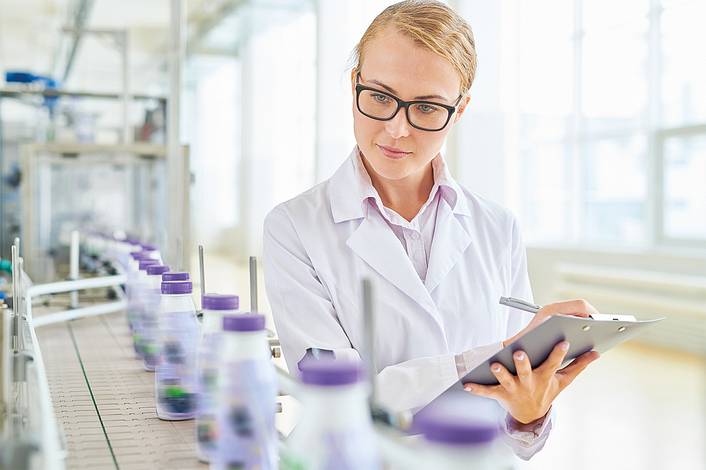 Eine Frau im Laborkittel mit Klemmbrett überprüft Flaschen auf einem Förderband.