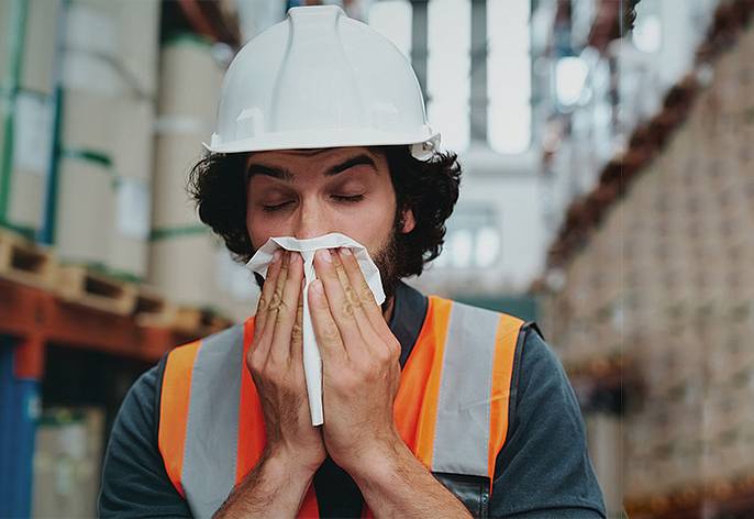An employee wears a high-visibility vest and hard hat and blows his nose in a logistics hall.