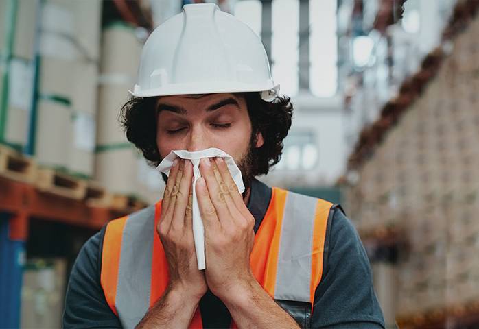 An employee wears a high-visibility vest and hard hat and blows his nose in a logistics hall.