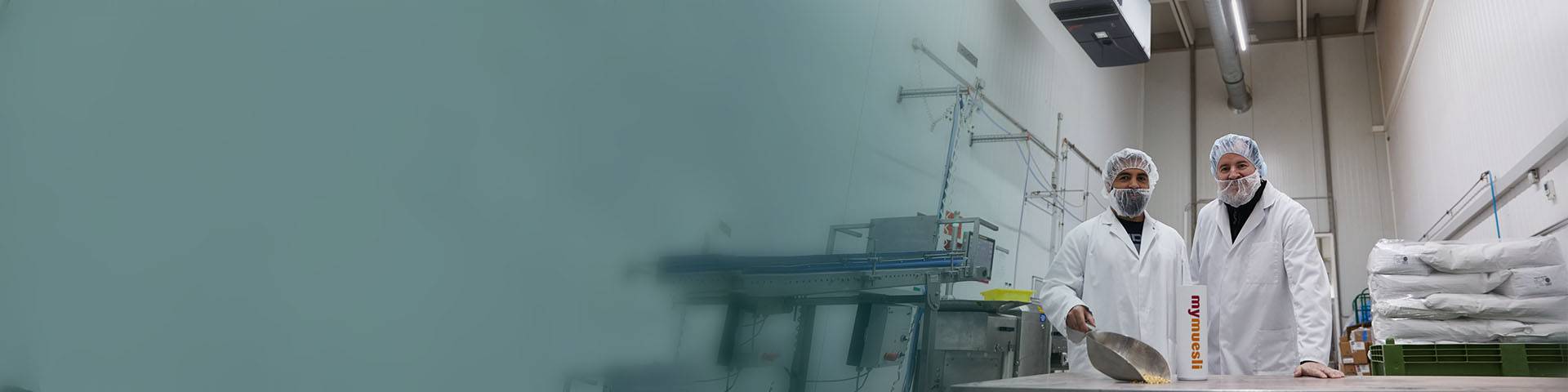 Mitarbeiter in Schutzkleidung mit Haar- und Barthaube lächeln in die Kamera und präsentieren Müsli.