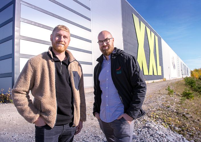 Two men stand in front of XXL Sports & Vildmark and smile into the camera.