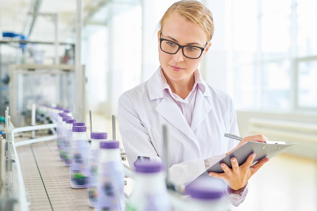 On a conveyor belt, a woman in a lab coat is checking bottles with a clipboard.
